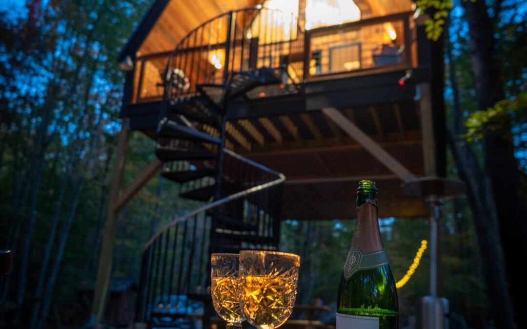Evening photo of a glamping experience with champagne in the foreground and a luxury treehouse in the background
